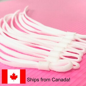 May include: A selection of white elastic cords with adjustable loops, possibly for face masks, presented on a pink surface. The cords are made of a soft, flexible material.