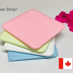 May include: Four pastel-coloured square plastic lids are stacked: pink, pale green, white, and blue. The text "Free Strap!" is visible. A cluster of bright pink flowers is to the right. The Canadian flag is in the lower right corner.