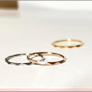 May include: Three delicate rings in silver, rose gold, and gold. Each ring features a faceted design, creating a geometric pattern around the band. The rings are displayed on a white surface, showcasing their minimalist style.