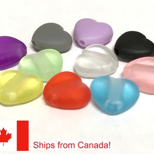 May include: A selection of heart-shaped beads in a range of colours: purple, grey, green, white, yellow, red, blue, pink, and black. Each bead has a hole for threading. The beads are arranged on a white surface.