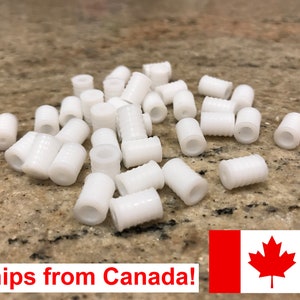 May include: A collection of small, white plastic cylindrical beads with a ridged exterior. The beads are scattered on a speckled, light brown surface. The image showcases a close-up view of the beads.