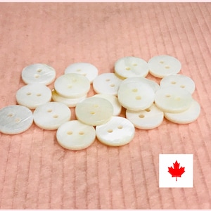 May include: A collection of round, off-white buttons with two holes each, scattered on a textured pink surface. The buttons have a pearlescent sheen, suggesting a mother-of-pearl material. The image includes a Canadian flag.