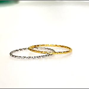 May include: Two thin rings, one silver and one gold, are shown. Both rings have a textured, faceted surface. The gold ring is positioned slightly behind the silver ring. The background is plain white.
