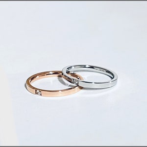 May include: Two thin rings, one rose gold and one silver, are displayed on a white surface. The rose gold ring has a small square gemstone. The silver ring also has a small square gemstone. Both rings are simple and elegant.