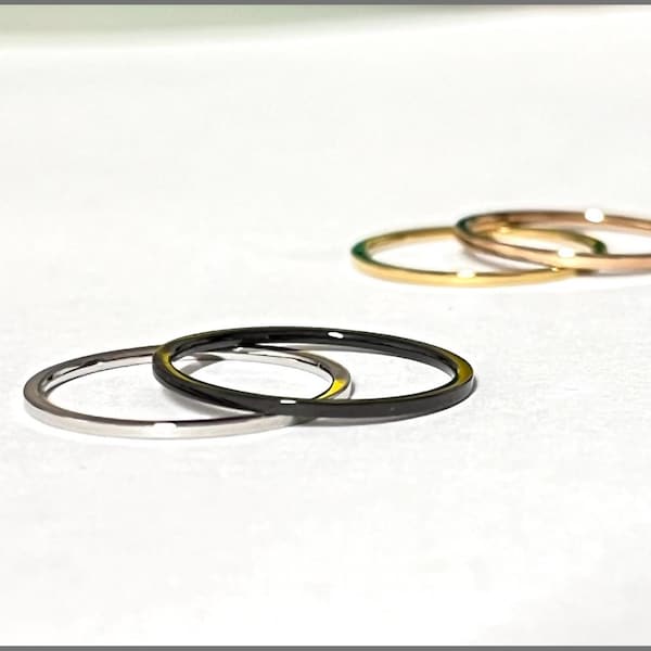 May include: A selection of thin, minimalist rings in different metallic finishes. The rings include silver, black, gold, and rose gold, each with a smooth, polished surface. The rings are displayed on a white background, highlighting their simple elegance.