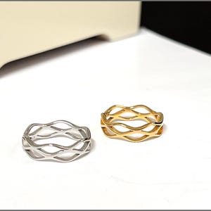 Peut inclure: Deux bagues avec un motif ondulé. Une bague est de couleur argentée et l'autre est de couleur dorée. Les bagues sont posées sur une surface blanche. Les bagues ont un motif ondulé continu.
