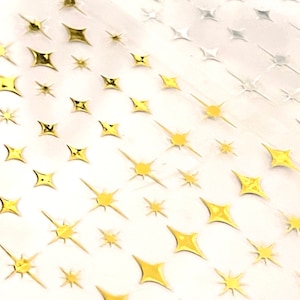 Op de afbeelding: Close-up van een vel met goud en zilverkleurige stervormige stickers. De stickers vari&euml;ren in grootte en ontwerp, sommige hebben puntige stralen en andere zijn compacter. De sterren zijn gerangschikt in een herhalend patroon.