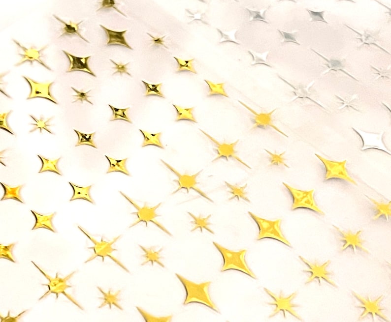 Op de afbeelding: Close-up van een vel met goud en zilverkleurige stervormige stickers. De stickers vari&euml;ren in grootte en ontwerp, sommige hebben puntige stralen en andere zijn compacter. De sterren zijn gerangschikt in een herhalend patroon.