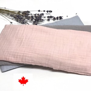 May include: Three rectangular fabric items in pink, grey, and blue are stacked. Dried lavender sprigs are placed above the fabrics. The pink fabric is in the foreground. The Canadian flag is in the bottom right corner.