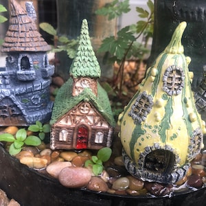 May include: Three miniature fairy houses made of painted ceramic. A grey stone tower with a pointed roof, a green church with a pointed roof, and a yellow gourd-shaped house with green accents.