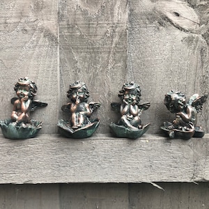 May include: Four small, bronze-coloured cherub figurines, each sitting on a leaf-shaped base. The cherubs are all wearing wings and have a thoughtful expression on their faces.