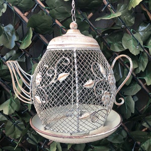 May include: A white metal bird feeder shaped like a teapot with a wire mesh design. The feeder has a small plate underneath for catching seeds.
