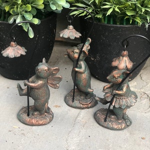 May include: Three bronze-coloured fairy figurines holding black poles with flower-shaped lights. The fairies have wings, skirts, and mouse-like features. The figurines are on a stone surface, with green plants in black pots in the background.