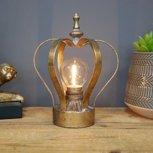 May include: A gold metal crown shaped table lamp with a light bulb inside. The lamp is on a wooden surface.