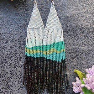 May include: Handmade beaded earrings with a triangular design. The earrings feature a gradient of colors, starting with white at the top, transitioning to shades of green and turquoise, and ending with a fringe of dark brown beads. The earrings hang from silver-colored hooks.
