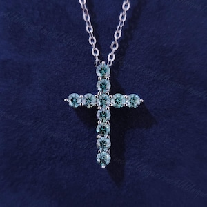 Könnte beinhalten: Eine silberfarbene Kreuz-Anhänger-Halskette mit einer filigranen Kette. Das Kreuz ist mit mehreren runden, hell türkis farbenen Edelsteinen besetzt. Die Halskette wird vor einem dunkelblauen Hintergrund präsentiert, der die Details des Schmucks hervorhebt.