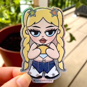 May include: A holographic sticker featuring a cartoon woman with blonde pigtails and star hair clips, holding a taco. She wears a denim outfit with white star accents. The sticker has a glittery border and the text "@fromelisabethshin".