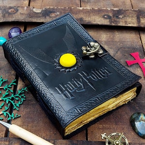 Peut inclure: Un journal en cuir noir avec un fermoir doré et les mots "Harry Potter" gravés sur la couverture. Le journal a un motif circulaire jaune avec un symbole ailé sur le devant.