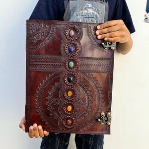 K&ouml;nnte beinhalten: Ein gro&szlig;es, braunes Lederjournal mit aufwendigen gepr&auml;gten Designs und sieben farbigen Edelsteinakzenten. Das Journal hat einen Verschluss und wird von einer Person gehalten. Das Cover hat ein kreisf&ouml;rmiges und geometrisches Muster.