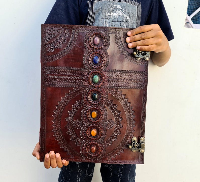 K&ouml;nnte beinhalten: Ein gro&szlig;es, braunes Lederjournal mit aufwendigen gepr&auml;gten Designs und sieben farbigen Edelsteinakzenten. Das Journal hat einen Verschluss und wird von einer Person gehalten. Das Cover hat ein kreisf&ouml;rmiges und geometrisches Muster.