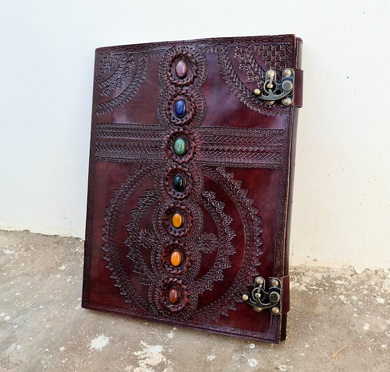 K&ouml;nnte beinhalten: Ein dunkelbraunes Lederjournal mit aufwendigen gepr&auml;gten Designs und einer Reihe farbiger Edelsteine in der Mitte. Das Journal hat einen Metallverschluss und detaillierte Muster. Die Abdeckung hat eine satte, rotbraune Farbe.