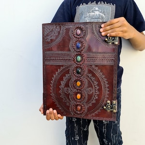 K&ouml;nnte beinhalten: Ein gro&szlig;es, dunkelbraunes Lederjournal mit aufwendigen gepr&auml;gten Designs und farbigen Edelsteinakzenten. Das Journal verf&uuml;gt &uuml;ber eine Reihe von sieben Edelsteinen in der Mitte und dekorative Metallverschl&uuml;sse. Das Cover ist mit detaillierten Mustern verziert.