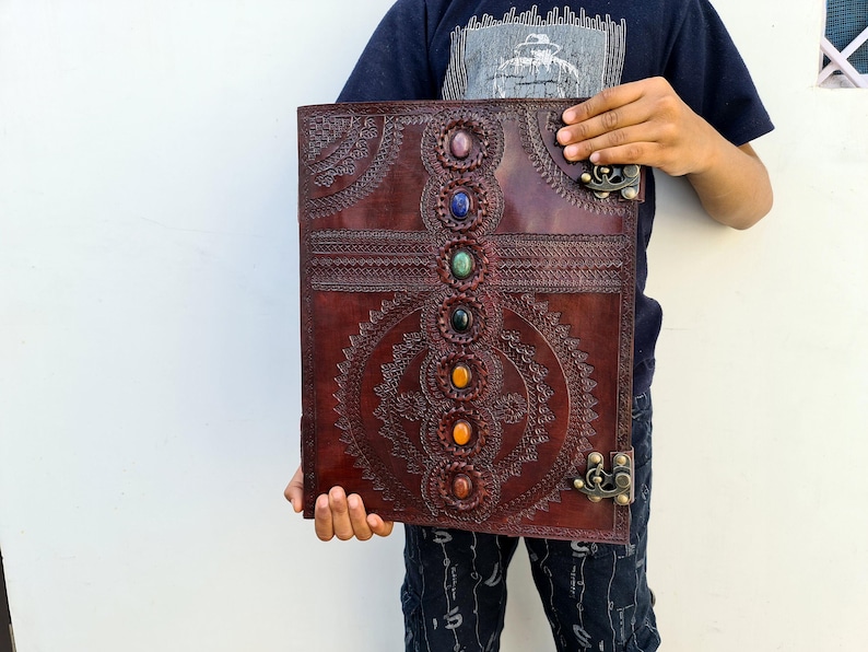 K&ouml;nnte beinhalten: Ein gro&szlig;es, dunkelbraunes Lederjournal mit aufwendigen gepr&auml;gten Designs und farbigen Edelsteinakzenten. Das Journal verf&uuml;gt &uuml;ber eine Reihe von sieben Edelsteinen in der Mitte und dekorative Metallverschl&uuml;sse. Das Cover ist mit detaillierten Mustern verziert.