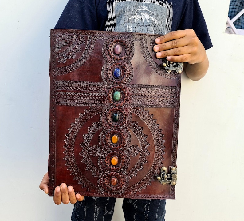 K&ouml;nnte beinhalten: Ein gro&szlig;es, dunkelbraunes Lederjournal mit aufwendigen gepr&auml;gten Designs und einer Reihe farbiger Edelsteine in der Mitte. Das Journal hat einen Metallverschluss und wird offen gehalten, um die detaillierte Handwerkskunst zu zeigen.