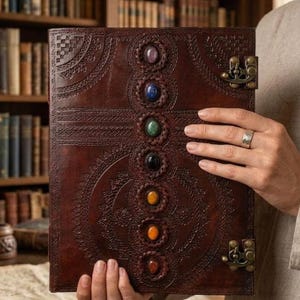 K&ouml;nnte beinhalten: Ein braunes Lederjournal mit aufwendigen gepr&auml;gten Designs und sieben farbigen Edelsteinen, die vertikal ausgerichtet sind. Das Journal hat einen Metallverschluss und wird offen gehalten, was die detaillierte Handwerkskunst und das spirituelle Thema zeigt.