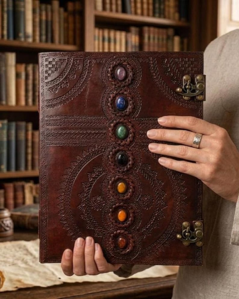 K&ouml;nnte beinhalten: Ein braunes Lederjournal mit aufwendigen gepr&auml;gten Designs und sieben farbigen Edelsteinen, die vertikal ausgerichtet sind. Das Journal hat einen Metallverschluss und wird offen gehalten, was die detaillierte Handwerkskunst und das spirituelle Thema zeigt.