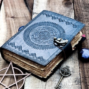 May include: A dark blue leather-bound journal with an intricate embossed design and a metal clasp. The journal is open to reveal aged, yellowed pages. A pentagram and a key are also visible in the image.