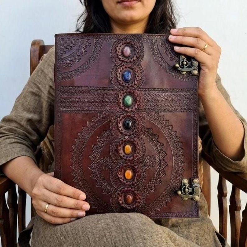 K&ouml;nnte beinhalten: Ein braunes Lederjournal mit einem detaillierten gepr&auml;gten Design und einer Reihe farbiger Edelsteine in der Mitte. Das Journal hat einen Verschluss und wird offen gehalten, was die komplizierte Handwerkskunst und das spirituelle Thema zeigt.