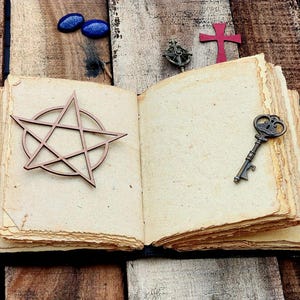 Może przedstawiać: Otwarty dziennik w stylu antycznym z brązowymi okładkami sk&oacute;rzanymi i postarzanymi papierowymi stronami. Na otwartych stronach umieszczono drewniany pentagram, mały klucz i czerwony krzyż.