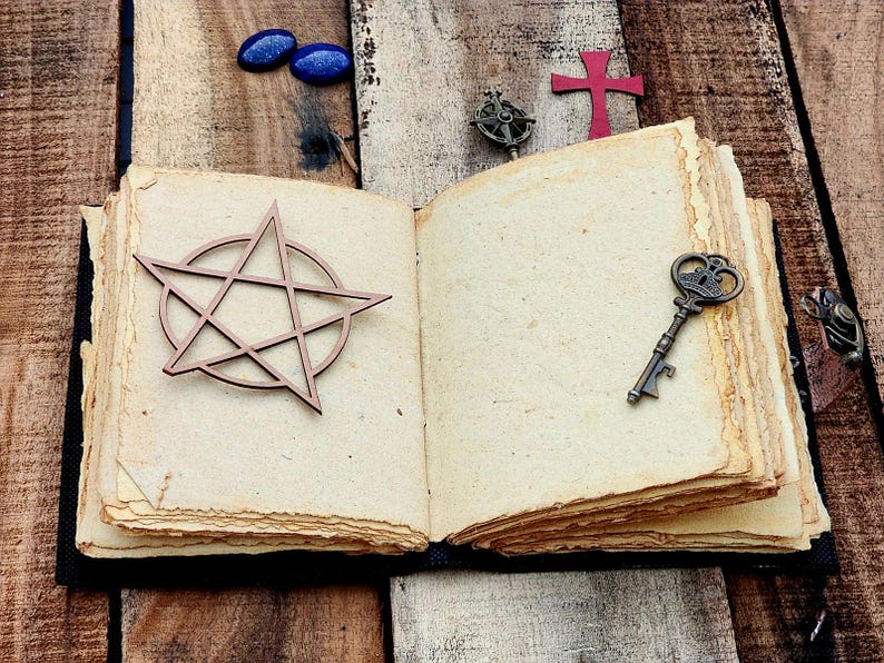 Może przedstawiać: Otwarty dziennik w stylu antycznym z brązowymi okładkami sk&oacute;rzanymi i postarzanymi papierowymi stronami. Na otwartych stronach umieszczono drewniany pentagram, mały klucz i czerwony krzyż.