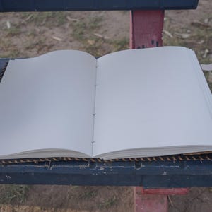K&ouml;nnte beinhalten: Ein offenes Notizbuch mit leeren wei&szlig;en Seiten. Das Notizbuch ist mit dunklem Seil gebunden und hat Lederriemen mit Metallverschl&uuml;ssen. Das Notizbuch liegt auf einer dunkelblauen Oberfl&auml;che mit einem gewebten Rand.
