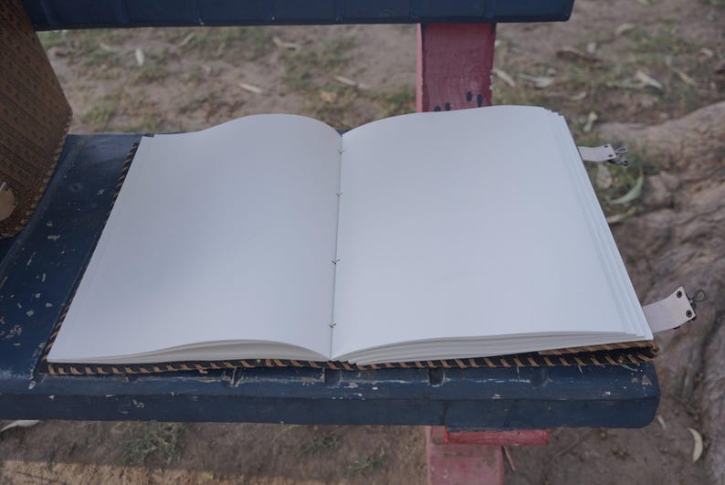 K&ouml;nnte beinhalten: Ein offenes Notizbuch mit leeren wei&szlig;en Seiten. Das Notizbuch ist mit dunklem Seil gebunden und hat Lederriemen mit Metallverschl&uuml;ssen. Das Notizbuch liegt auf einer dunkelblauen Oberfl&auml;che mit einem gewebten Rand.