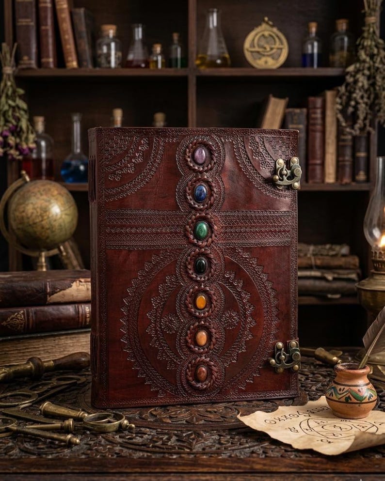 K&ouml;nnte beinhalten: Ein gro&szlig;es, braunes Lederjournal mit gepr&auml;gten Mustern und sieben farbigen Edelsteinen. Das Journal hat einen Metallverschluss und wird auf einem Holztisch mit anderen antiken Gegenst&auml;nden, darunter B&uuml;cher und ein Globus, ausgestellt.