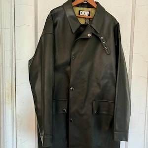 May include: A black jacket with a smooth finish, featuring a pointed collar, a front button closure, and two lower flap pockets. The interior lining is a light olive green color with a black DKNY brand label visible at the neckline.