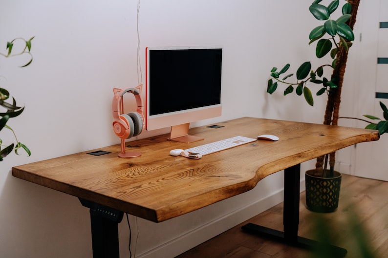 K&ouml;nnte beinhalten: Ein Holzschreibtisch mit einer Live-Edge-Oberfl&auml;che und schwarzen verstellbaren Beinen. Auf dem Schreibtisch befinden sich ein Computermonitor, eine Tastatur, eine Maus und ein rosa Kopfh&ouml;rer.