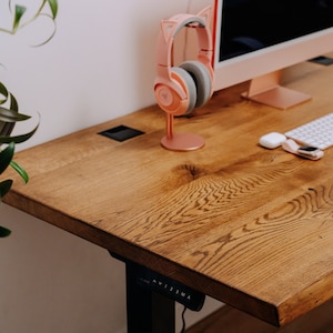 Pode incluir: Uma mesa de madeira com acabamento marrom claro. A mesa tem uma tomada de energia integrada e um mecanismo de altura ajustável preto com botões etiquetados "A, V, 1, 2, 3, M, A". Um fone de ouvido rosa com orelhas de gato está em um suporte na mesa.