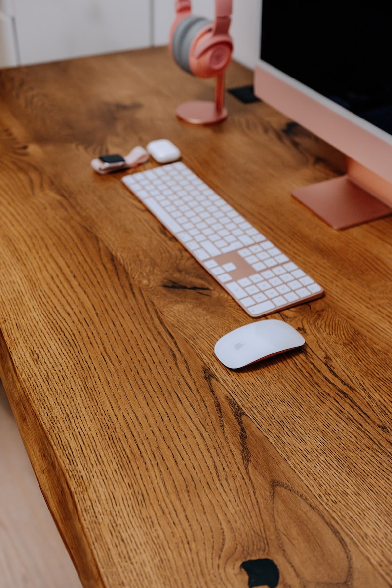K&ouml;nnte beinhalten: Ein Holzschreibtisch mit einer wei&szlig;en kabellosen Tastatur, einer wei&szlig;en kabellosen Maus und einem rosa Kopfh&ouml;rerst&auml;nder mit rosa Kopfh&ouml;rern.