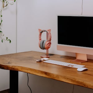 K&ouml;nnte beinhalten: Ein ros&eacute;goldener Computermonitor mit schwarzem Bildschirm steht auf einem Holztisch. Ein rosa Kopfh&ouml;rer mit Katzenohren steht auf einem St&auml;nder neben dem Monitor. Eine wei&szlig;e kabellose Tastatur und Maus befinden sich auf dem Tisch.