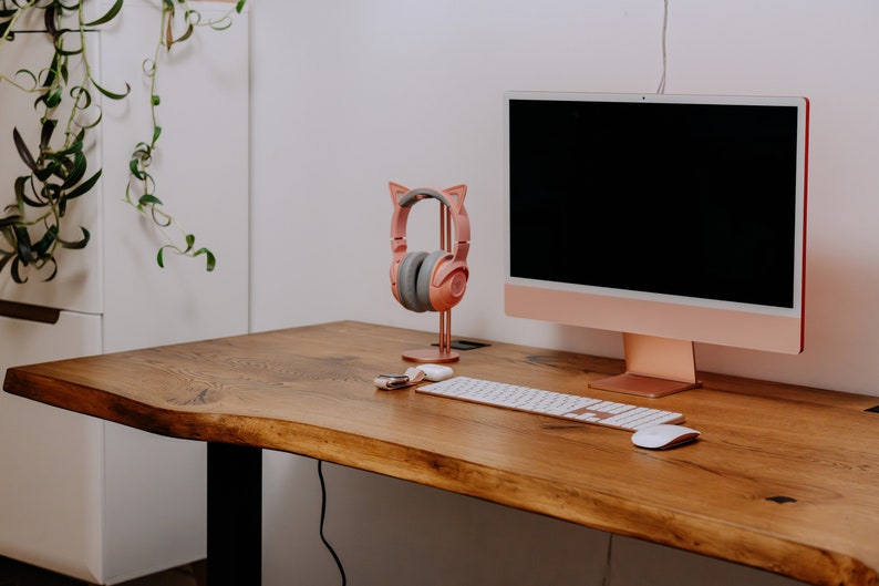 K&ouml;nnte beinhalten: Ein ros&eacute;goldener Computermonitor mit schwarzem Bildschirm steht auf einem Holztisch. Ein rosa Kopfh&ouml;rer mit Katzenohren steht auf einem St&auml;nder neben dem Monitor. Eine wei&szlig;e kabellose Tastatur und Maus befinden sich auf dem Tisch.