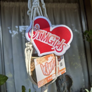 May include: A red heart-shaped patch with the text "OTM Girls" in white script, framed by a white border. Below, a card with the word "Vivi" and floral designs is visible, all suspended in a macrame plant hanger.
