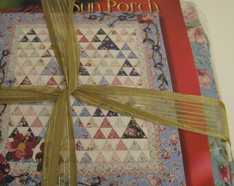 Könnte beinhalten: Ein Stapel Bücher, gebunden mit einem goldenen Band. Das oberste Buch zeigt ein Steppmuster mit dreieckigen Formen in Blau, Rosa und Weiß. Der Titel des Buches lautet "Sun Porch". Andere Bücher haben florale Muster.