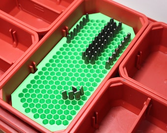 May include: A red plastic storage organizer tray featuring a bright green hexagonal grid insert. The insert holds a collection of dark metal screwdriver bits arranged in rows, with a few silver-colored flat-head bits positioned separately.