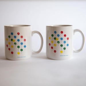 May include: Two white ceramic mugs with a design of colorful balloons. The mugs have handles and the text "Simply Balloon Company" on the bottom.