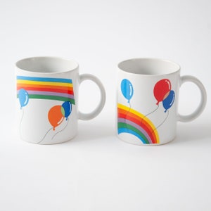 May include: Two white ceramic mugs with rainbow designs and colorful balloon illustrations. The mugs have handles and are perfect for hot beverages.