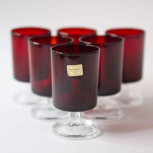 May include: Six red glass tumblers with a clear glass stem. The tumblers are all the same size and shape. The tumblers have a label that says "Luminarc" on them.