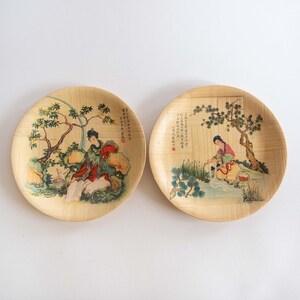 May include: Two round bamboo plates with hand-painted scenes of figures in traditional clothing. The plates feature detailed illustrations with trees, rocks, and flowing water, showcasing a classic Asian art style. The plates are a light tan color.